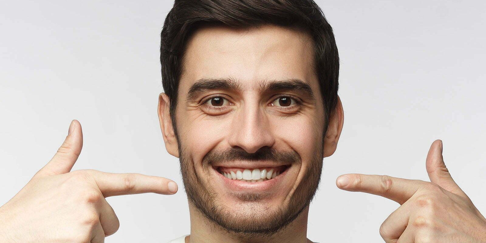 Portrait of smiling young man pointing to his perfect natural white teeth after whitening treatment. Dental clinic ads concept