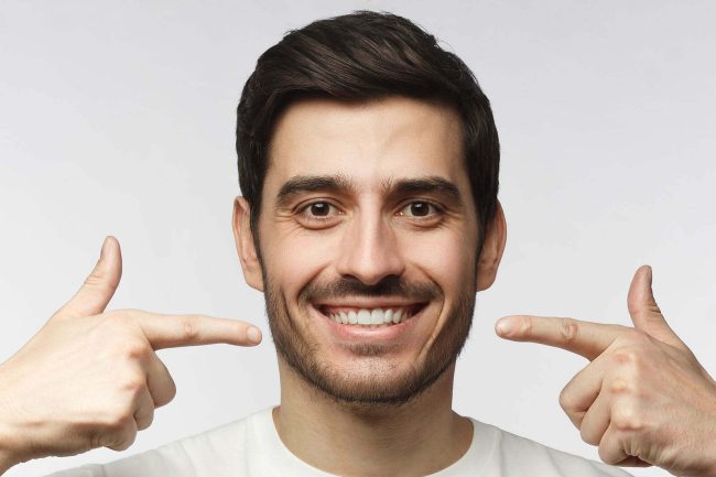 Portrait of smiling young man pointing to his perfect natural white teeth after whitening treatment. Dental clinic ads concept